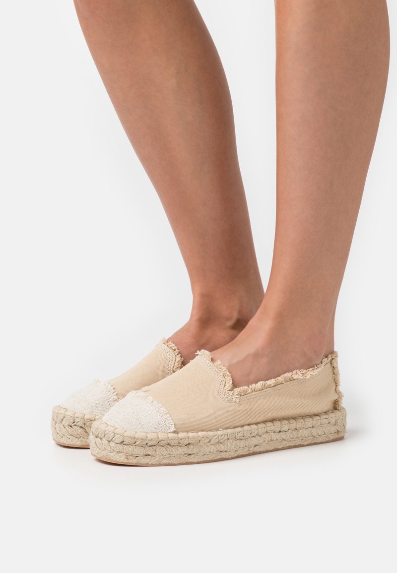 Pieds portant des espadrilles en toile beige avec des bords effilochés et des semelles en jute tressé sur fond blanc.