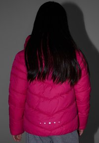Personne aux longs cheveux foncés portant une doudoune rose vif, debout de dos sur un fond gris uni.