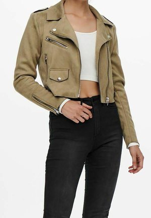 Femme portant une veste biker en daim beige courte avec des fermetures éclair et des boutons argentés, un crop top blanc et un pantalon noir taille haute.