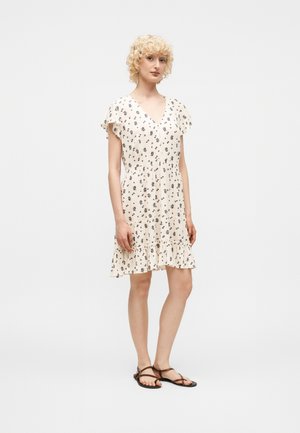 Kobieta z krótkimi kręconymi blond włosami, ubrana w białą kwiecistą sukienkę z krótkim rękawem i brązowe płaskie sandały, stoi na jednolitym tle.