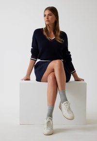 Vrouw die op een witte blok zit, in een marineblauwe trui, marineblauwe rok, grijze sokken en witte sneakers, kijkend naar de zijkant in een neutrale omgeving.