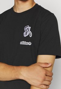 Svart bomulls-t-shirt med ett vitt broderat Goofy-motiv på vänster bröst, med en kontrasterande Adidas-logotyp nedanför.