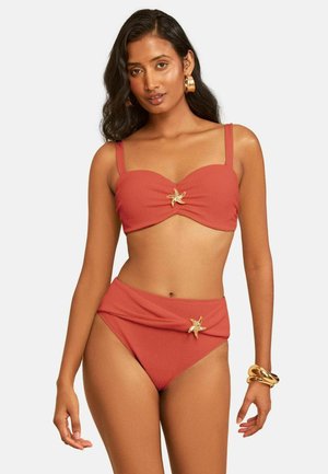 Kvinde iført rød tekstureret bikini med guld søstjerne-accenter på top og bund, guld øreringe i ringe og chunky armbånd på højre håndled.
