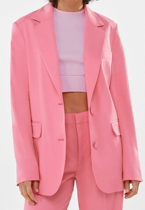 Persoon draagt een felroze oversized blazer en bijpassende broek met daaronder een lichtpaars cropped geribd topje, tegen een effen achtergrond.