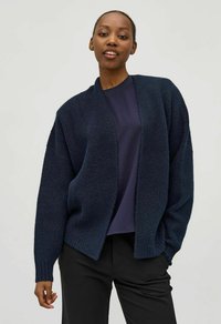 Cárdigan de punto azul marino con frente abierto, textura acanalada, hombros caídos y mangas largas, llevado sobre una camiseta oscura y pantalones negros.