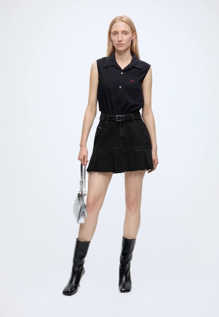 Femme portant un haut noir sans manches avec col et une jupe noire avec ceinture, tenant un petit sac à main argenté et portant des bottines noires.