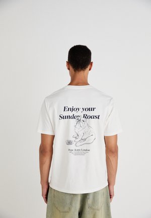 Weißes Baumwoll-T-Shirt mit kurzen Ärmeln, das eine Bulldoggen-Illustration und den Text "Genieße deinen Sonntag Braten" auf dem Rücken zeigt.