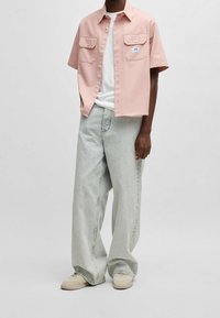 Chemise rose à manches courtes avec poches poitrine, t-shirt blanc en dessous, associée à un jean bleu clair décontracté et des baskets beiges.