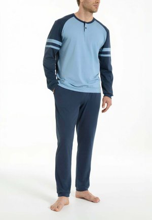 Uomo in piedi a piedi nudi, indossa una maglia blu a maniche lunghe con bottoni e pantaloni blu scuro, mani nelle tasche, rivolto leggermente verso destra.