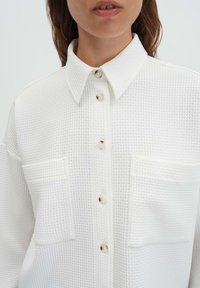 Camicia bianca testurizzata con scollo a collo, con chiusura a bottoni e due tasche frontali. Il tessuto presenta un motivo a diamante sottile.