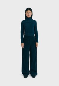 Mørkeblå, tekstureret turtleneck-bluse parret med høj talje og brede bukser i matchende stof, båret med sorte sko på en lys baggrund.