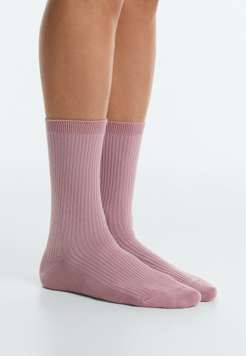 Jambes portant des chaussettes côtelées mi-mollet de couleur rose poudré, debout sur une surface blanche sur fond clair.