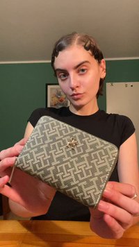 Novčanik na zatvaranje s teksturiranom sivoj tkanini s geometrijskim uzorkom. Sadrži zlatni logotip i zaobljene rubove.