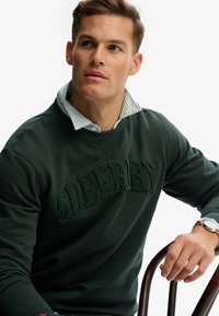 Felpa verde scuro con la scritta in rilievo "SUPERDRY" sul davanti, indossata sopra una camicia a righe bianche e azzurre, con una finitura texturizzata.