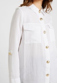 Camisa blanca, ligera, con cuello, dos bolsillos en el pecho y botones en tono dorado. Presenta mangas con opción de vuelta y una textura semi-translúcida.