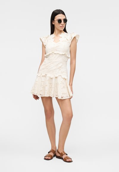 Γυναίκα που φοράει φόρεμα από κρεμ δαντέλα με βολάν στις άκρες, καφέ σανδάλια και στρογγυλά μαύρα γυαλιά ηλίου, στέκεται σε απλό φόντο.