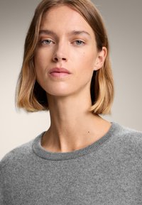 Grauer Strickpullover mit rundem Ausschnitt, weicher Textur und geripptem Saum. Das Model hat schulterlanges Haar mit einem natürlichen Finish.