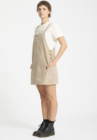 Beige corduroy salopette jurk met een rechte silhouet, voorzien van zijzakken, knopen op de banden, en gecombineerd met een wit graphic t-shirt.