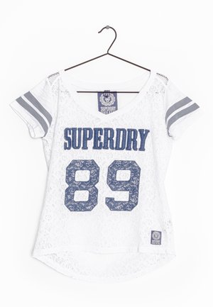 Chemise blanche en dentelle à manches courtes avec texte bleu "SUPERDRY 89", deux bandes grises sur chaque manche, suspendue à un cintre noir.