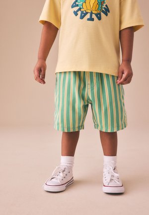 Kind trägt gelbes kurzärmeliges Shirt mit Cartoon, grün-gelb gestreifte Shorts, weiße Socken und weiße Sneaker und steht auf beigem Boden.