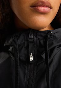 Czarna nylonowa kurtka z zamkiem błyskawicznym z przodu, kapturem z regulowanym sznurkiem i metką z logo. Gładka faktura z widocznymi detalami szwów.