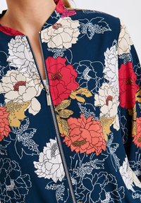 Chaqueta con cremallera y estampado floral, con fondo azul marino, grandes flores de peonía y hojas en rojo, blanco y amarillo, con textura de tela suave y cuello alto.