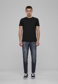Fekete pamut póló, slim-fit, sötétkék farmernadrággal párosítva, amelyen szakított részletek és elhasználódott területek találhatók. Fekete-fehér sneaker cipők teszik teljessé a megjelenést.