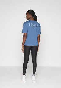Plava pamučna majica kratkih rukava s natpisima "TRAIN" i "Pretty Tough" na leđima. Nosena s crnim tajicama i bijelim sportskim cipelama.