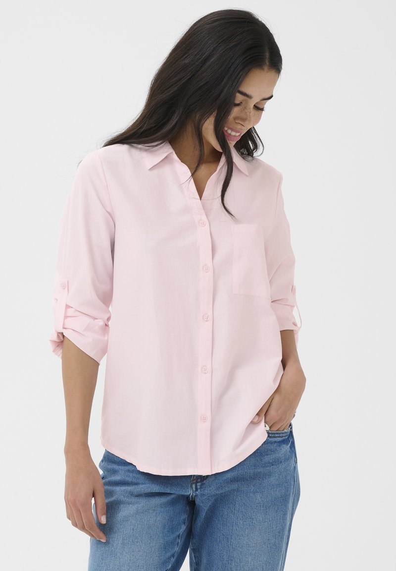 Femme aux longs cheveux bruns portant une chemise rose clair à boutons avec les manches retroussées et un jean bleu, souriante et regardant vers le bas.