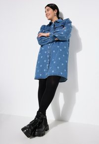 Jeanshemdkleid in Hellblau mit Herzmustern, langen Ärmeln, Knopfleiste, kombiniert mit schwarzen klobigen Stiefeln mit Absatz und schwarzen Strumpfhosen.