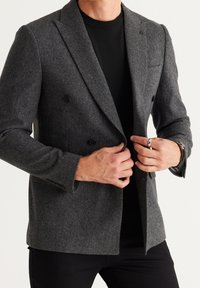 Donkergrijze wollen blazer met een double-breasted ontwerp, met inkeping kraag, en twee zwarte knopen; gedragen over een zwart shirt met een ronde hals.