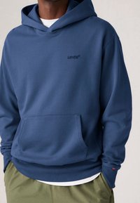 Modrá bavlněná mikina s kapucí, přední kapsou, žebrovanými manžetami a spodním lemem, na hrudi je vyšitý logo "Levi's" a je vrstvená přes bílé tričko.