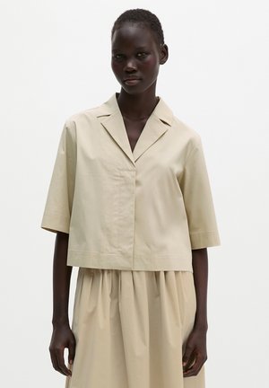 Kvinde iført en løs beige kortærmet skjorte med krave og matchende plisseret nederdel mod en ensfarvet lys baggrund.