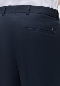 Pantalon bleu marine foncé avec une poche passepoilée à l’arrière et un petit détail de logo métallique, porté avec une chemise blanche rentrée.