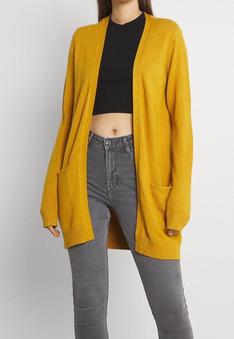 Personne portant un cardigan ouvert jaune moutarde sur un top court noir et un jean gris taille haute sur fond uni.