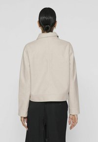 Chaqueta de mezcla de lana beige clara con cuello, diseño corto y mangas largas. Presenta costuras visibles y un estilo minimalista.
