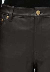 Črne hlače iz umetnega usnja z zlatim okovjem, vidnim šivanjem in klasičnim oblikovanjem s petimi žepi ter gladko teksturo.
