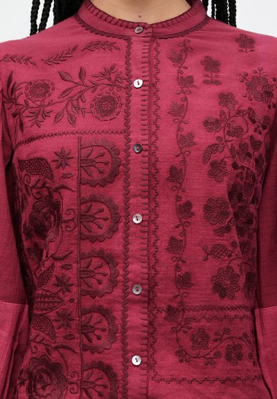 Vīna sarkana krekla virsdaļa ar smalku tumšu ziedu izšuvumu, teksturētu audumu un apaļu apkakli. Ir četri pogas un detalizētas šuves.