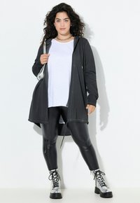 Cardigan lungo nero con cappuccio, abbinato a una maglietta bianca e leggings neri lucidi, completato da stivali lacciati argento. Texture liscia.