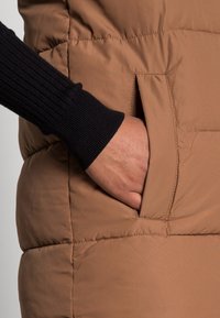 Hand med mörk ärm som är insatt i sidofickan på en brun vadderad jacka eller väst.