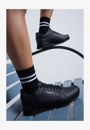 Czarne sneakersy Puma mają gładki materiał syntetyczny, okrągły czubek, fakturowane detale oraz logo z boku, w połączeniu z czarno-białymi skarpetami w paski.