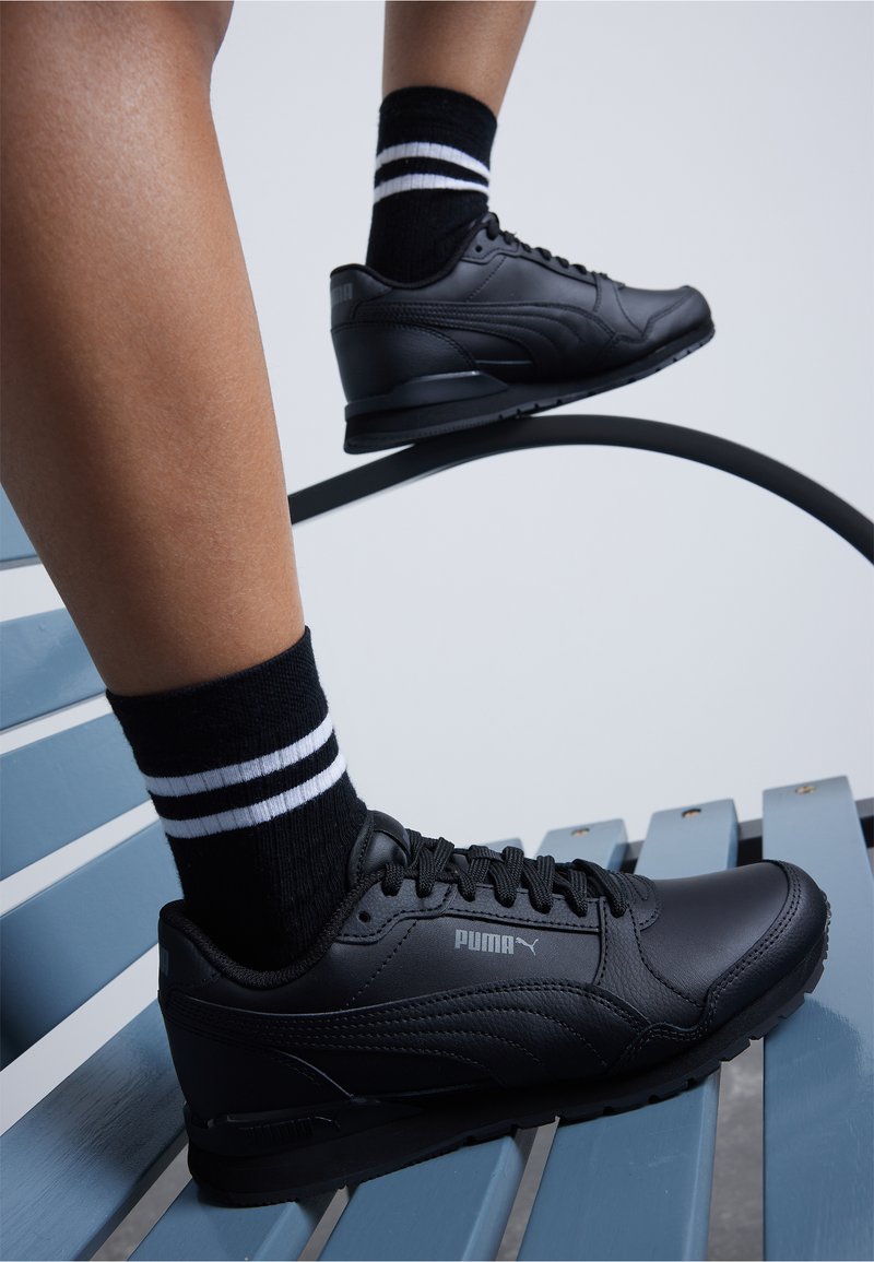 Czarne sneakersy Puma mają gładki materiał syntetyczny, okrągły czubek, fakturowane detale oraz logo z boku, w połączeniu z czarno-białymi skarpetami w paski.