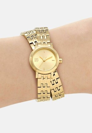 Orologio da polso DKNY tono oro con quadrante rotondo e cinturino a doppio anello, indossato su un polso dalla pelle chiara che segna le 4:20.