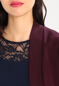 Kvinnas nedre ansikte och hals klädd i mörk spetsöverdel och burgundy cardigan, med rosa läppstift och mörkt hår synligt.