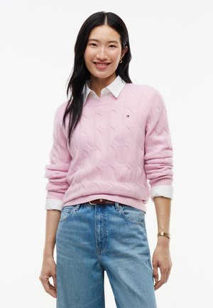 Kvinde smilende, iført en lyserød kabelstrikket sweater over en hvid skjorte med krave, blå jeans og et gyldent armbåndsur.