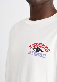 White short-sleeve T-shirt, featuring multicolored graphic text "VOLCOM" in red and "STONE" in purple, with a black eye design. Cotton material.