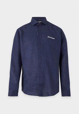 Homme portant une chemise bleu marine à boutons sur un T-shirt blanc, un pantalon bleu marine assorti et des baskets blanches, debout devant un fond uni.