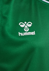 Zielony materiał sportowy z białym logo "hummel" w stylizowanym kształcie pszczoły, gładka tekstura i minimalistyczne akcenty.