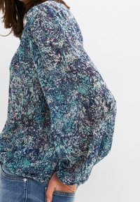 Blouse à manches longues avec un motif abstrait bleu et vert, dotée d'épaules froncées et d'une coupe décontractée, associée à un jean en denim.