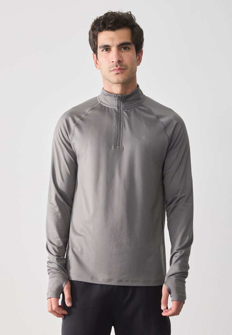 Grijze pullover met lange mouwen en rits tot een kwart, zachte textuur, sportieve pasvorm en duimgaten bij de manchetten, gedragen met donkere broek.
